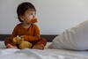 Kleinkind mit Stofftier und Schnuller im Mund sitzt auf dem Bett 