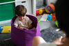Kleinkind sitzt in einem Spielzeugkorb und beschäftigt sich konzentriert mit einem Spielzeug, während die Mutter vor ihm sitzt.