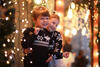 Zwei Kleinkinder umgeben von Weihnachtsbeleuchtung stehen hintereinander und freuen sich
