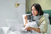 Kranke Frau sitzt mit Schal, warmer Decke und einer Tasse Tee im Bett