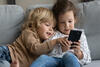 Zwei Kinder sitzen auf der Couch und beschäftigen sich mit einem Smartphone