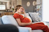 Schwangere sitzt mit geschlossenen Augen auf der Couch und hält sich mit einer Hand den Bauch und mit der anderen den Kopf