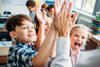 Grundschulkinder stehen hintereinander im Klassenraum und geben fröhlich ein High Five mit dem Lehrpersonal