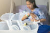 Manuelle Milchpumpen und eine Spritze gefüllt mit Kolostrum, im Hintergrund stillt Mutter ihr Neugeborenes im Klinikbett
