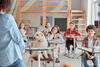 Schüler*innen sitzen im KLassenraum an Einzeltischen, dazwischen sitzt ein Labradoodle ebenfalls auf einem Stuhl an einem Tisch