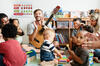 Erzieher sitzt mit Gitarre und einer weiteren Erzieherin und Kindern im Kreis auf dem Fußboden, die auch auf Musikinstrumenten spielen