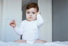 Baby sitzt auf dem Bett, hält eine Hand ans Ohr und guckt unglücklich in die Kamera