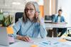 Grauhaarige Managerin sitzt am Arbeitsplatz mit Laptop und Haftnotizen